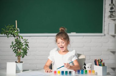 Çocuk kız sınıfta resim çiziyor, masada oturuyor, okulun karatahta arka planında eğleniyor. Çocukların yaratıcı eğitimi