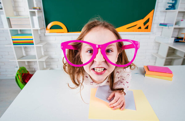 Funny angry child school girl with fun glasses in classroom. Crazy child grimacing in classroom near blackboard desk