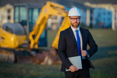 İnşaat mühendisi inşaat alanında çalışıyor. Olgun mühendis. İnşaat alanında takım elbiseli ve kasklı bir adam var. Orta yaşlı inşaat mühendisi kazıcının yanında duruyor.