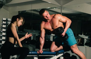 Kas yığını olan bir adamla kadın antrenör, kaslarını çalıştırıyor, dambıl kaldırıyor. Güç, güç ve sağlıklı yaşam tarzı, spor