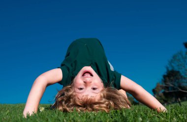 Mutlu çocuk yeşil çimenlerde baş aşağı oynuyor. Baharın bahçesinde komik bir çocuk. Çocuk günü. Mutlu tasasız çocukluk konsepti