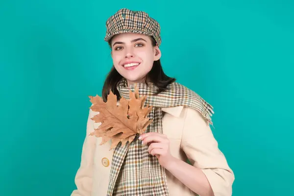 Hipster young woman in trendy autumn fall outfit stylish hat and scarf. Beauty girl hold autumn leaves near face, isolated on studio background. Portrait of autumnal woman with maple leaf