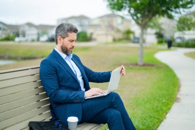Parktaki bankta oturan bir iş adamı. Adam dışarıda bilgisayarla uğraşıyor. Parktaki bankta oturup dizüstü bilgisayarla online iş yapan yakışıklı bir iş adamı. Takım elbiseli bir iş adamı parkta haber okuyor.
