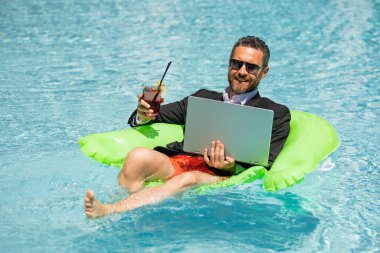 Takım elbiseli bir iş adamı yaz kokteyli içer ve havuzda dizüstü bilgisayar kullanır. Seyahat sektörü turizmi. Yazın bilardoda dizüstü bilgisayar kullanan bir ofis çalışanı. Yaz havuzunda başarılı iş adamı hayalleri