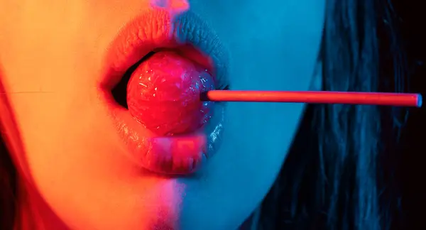 Lollipop in the mouth, close-up. Beautiful girl mouth with lolli pop. Glossy red woman lips with tongue. Mouth lick suck chupa chups on neon lights. Night club background