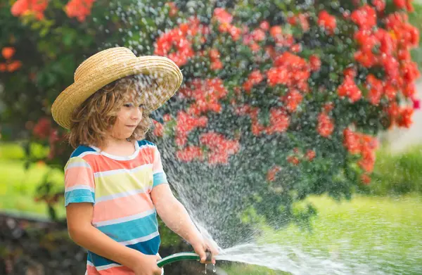 Happy kid boy pours water from a hose. Child watering flowers in garden. Home gardening