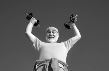 Yaşlı adam dambılları kaldırırken onun önünde ellerini tutuyor. Kıdemli adam mavi gökyüzünde egzersiz yapıyor. Spor merkezinde kıdemli sporcu.