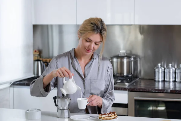 Young woman have breakfast, pouring milk in coffee. Woman at home in morning. Beautiful sensual female housewife in sexy pajama in kitchen cooking breakfast