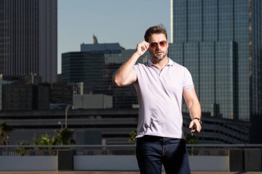 Sokakta polo tişörtlü yakışıklı bir genç. Dışarıda poz veren yakışıklı, kendine güvenen bir hippi modeli. Amerikan şehrinin arka planında seksi moda erkeği
