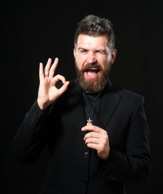 Beyaz arka plan üzerinde duran rahip üniforması giyen genç adam el hareketleriyle gülümsüyor ve mutlu yüzle parmaklarının arasından bakıyor.