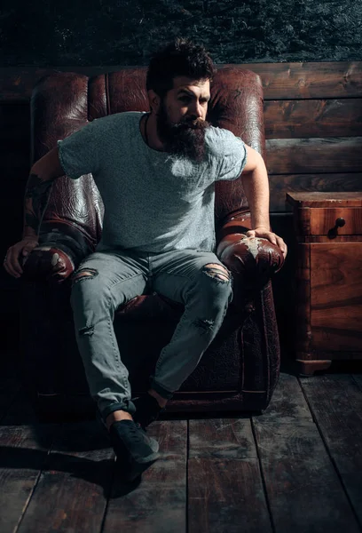 Handsome confident bearded man sitting in luxury leather chair and looks grumpy. Stylish attractive man with dramatic face. Bearded man wearing informal clothes and sitting in chair