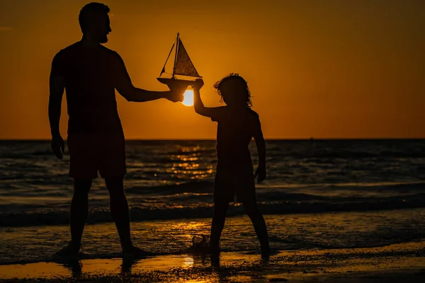 Son and father playing with toy boat in summer sea. Family dream on cruise liner. Travel, summer holiday. Journey trip. Dad and son playing with toy seailing boat on sunset sea. Fathers day