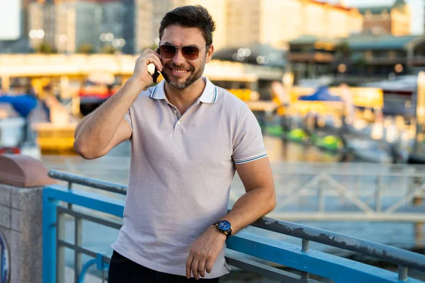 Portrait of handsome male model chatting on phone outdoor. Stylish man talking on phone dressed in polo. Fashion male posing on the street background. Urban style. Business phone conversation