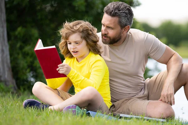 Father read a book with son in a park outdoors. Father and child son reading outdoor on green nature background. Dad with kid reading book together in the summer park. Family reading a book in nature