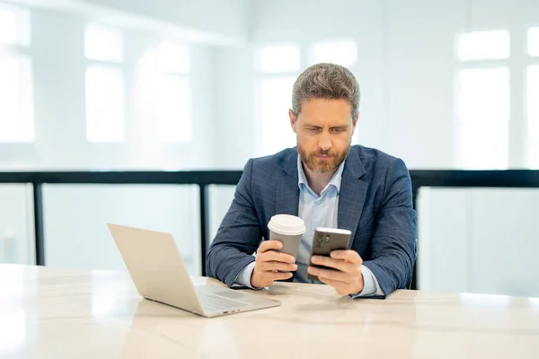 Business man in suit in office talk on phone. Office worker using phone, office call center. Man talk on phone work on laptop. Businessman have business call, talking on phone. Modern office interior