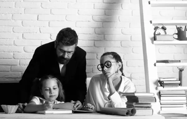 Portrait of father tutor and school kids daughters at school. Reading book and writing. Dad teaching schoolchild girls in classroom