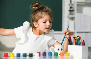 Çocuk kız sınıfta resim çiziyor, masada oturuyor, okulun karatahta arka planında eğleniyor. Erken eğitim