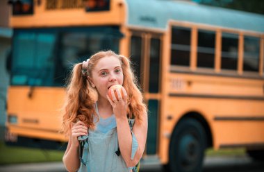 Eğitim: Gülümseyen öğrenci kız otobüse binmeye hazır. Okul otobüsünün önündeki kız elma yiyor.