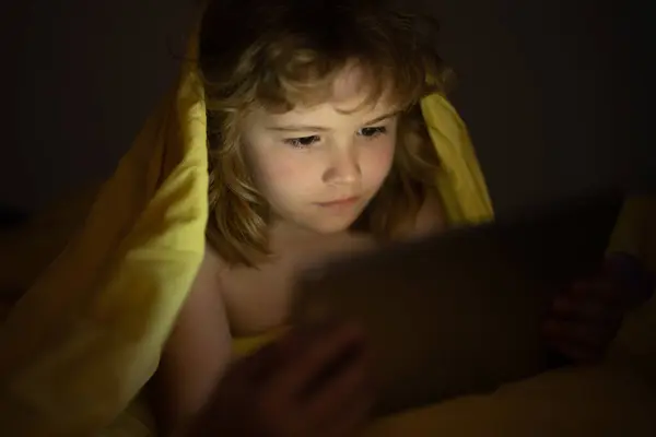 Kid watching tablet before sleeping. Child reads the e-book. Little boy looks at the screen of the tablet at hight. Kid boy playing tablet lying on a bed under blanket. Child social media addiction