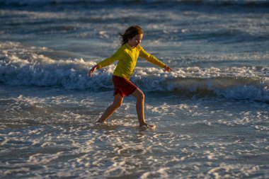 Yazın tropik plajlarda koşan ve oynayan bir çocuk. Çocuk koşmaktan zevk alıyor. Denizde yaz tatili. Çocuk gün batımında sahilde koşuyor. Çocuklar yaz tatilinde denizde koşarlar. Çocuk deniz suyunda koşar