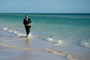Serbest iş, online iş. Yaz işi. Takım elbiseli iş adamı dizüstü bilgisayarı tutuyor ve denizde koşuyor. Suda dizüstü bilgisayarı olan takım elbiseli komik bir iş adamı. Uzaktan kumanda. Çılgın serbest çalışan.