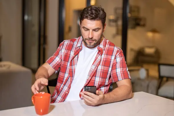 Portrait of man looking at smart phone at living room. Man is talking hold cup of coffe chatting on the smartphone. Thoughtful man using mobile phone at home. Middle-aged man using phone