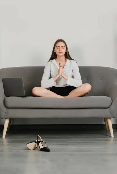 Meditation woman in a modern workplace doing yoga exercise on sofa at workplace in a modern office. Employee feel balance harmony relaxation. Calm accountant girl on sofa in office