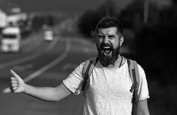 Auto stop travel. Hipster try to stop car and thumb up gesture. Man with cheerful face and beard travelling by hitchhiking with road on background. Travelling and hitch hiking concept