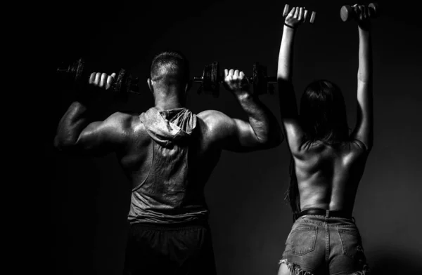 Sexy sport couple exercising with dumbbell. Muscular man with naked body, fitness woman with dumbbells on a dark background, back view. Slim sexy girl with strong muscular man workout
