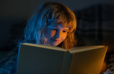 Çocuk karanlık odada masal okuyor. Kitap okuyan şirin bir çocuk. Açık kitabı olan bir çocuk. Ev okulu dersi. Evde kitap okuyan konsantre küçük çocuk.