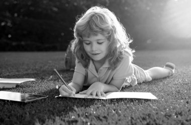 Not defterine kalem yazıp çimlere uzanan sevimli çocuk. Çocuk yaz parkında kitap okuyor.