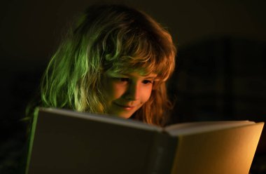 Çocuk masal okuyor, karanlık oda fotoğrafı. Çocuk yatakta sihirli bir kitap okuyor. Çocuk evde kitap okuyor.
