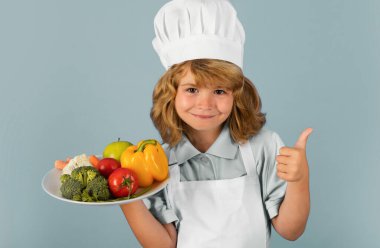 Çocuk fırın üniforması giyiyor ve aşçı şapkası takıyor. Mutfakta sebze hazırlıyor, stüdyo portresi yapıyor. Aşçılık, aşçılık ve çocuk maması konsepti.