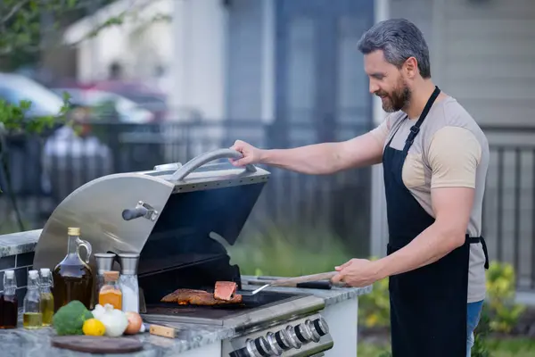 Men cooking on barbecue grill in yard. Cook at a barbecue grill preparing meat. Guy cooking meat on barbecue for summer family dinner at the backyard of the house