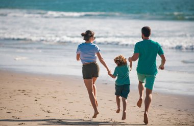 Sahilde koşan aile manzarası. İnsanlar yaz tatilinde eğleniyor. Baba, anne ve çocuk mavi denize karşı el ele tutuşuyorlar. Tatil seyahati kavramı