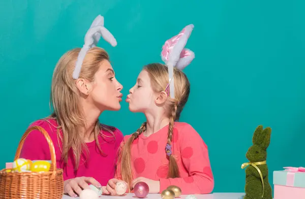 Attractive young woman with little cute girl are preparing for Easter. Mother and daughter wearing bunny ears are kissing spending time together before Easter while painting eggs