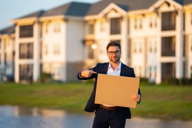 Açık havada poz veren boş afişi olan bir iş adamı. Takım elbiseli çekici bir adamın portresi boş boş bir poster tutuyor. Boş poster gösteren, işaret eden ve işaret eden bir adam.