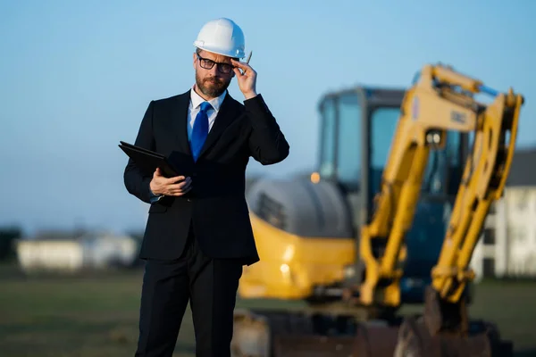Civil engineer worker at a construction site. Mature engineer worker. Man in suit and hardhat helmet at construction site. Middle aged head civil engineer worker standing outside near excavator