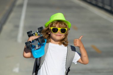 Parkta güneşli bir günde çocuk kaykayı tutuyor. Küçük kaykaycının açık hava portresi. Yaz parkında kaykay yapan bir çocuk, kaykay yapmayı öğreniyor. Başparmağı havada hayret bir çocuk, sokak