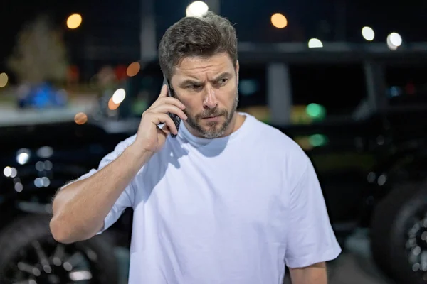 Angry man talking on phone near car on night urban street. Dangerous aggressive man talking on phone with serious face. Criminal city. Danger district. Aggressive angry man talking on phone outdoor