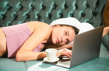 Kız dinleniyor, dizüstü bilgisayarla sohbet ediyor, lüks yatak odasında rahat bir kanepede kahve içiyor. Çekici seksi kadın otel odasında bornoz ve havlu giyerek rahatlıyor.