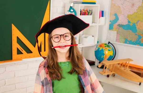 Portrait of funny schoolgirl with pen in mouth in graduation hat in classroom. Child studying