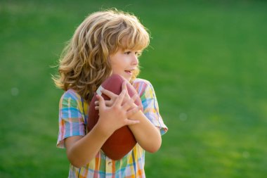 Ragbi toplu çocuk portresi. Çocuklar ve spor. Amerikan futbolu oynayan genç bir çocuk. Summer Park 'ta Amerikan futbolu oynarken elinde ragbi topu tutan çocuk.