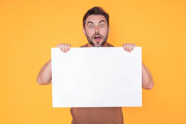 Yakışıklı, şaşkın bir adam elinde boş tahtayla stüdyoyu işaret ediyor. Kopyalama alanı olan boş tabela gösteren harika bir adam. Fikir ve teklif. Reklamınız. Boş reklam panosu