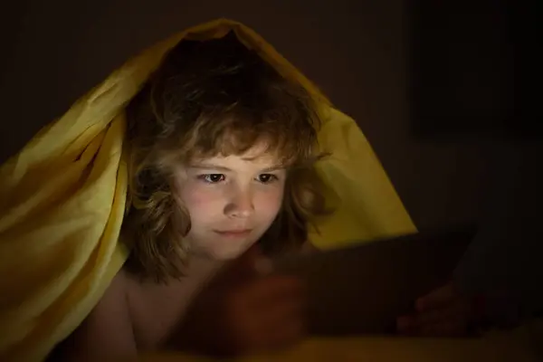 Kid watching tablet before sleeping. Child reads the e-book. Little boy looks at the screen of the tablet at hight. Kid boy playing tablet lying on a bed under blanket. Child social media addiction