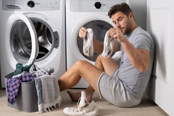 Middle aged single man with laundry basket and dirty clothes near washing machine at home. Millennial man putting clothes into washing machine. Displeased hispanic man wash clothes in washing machine