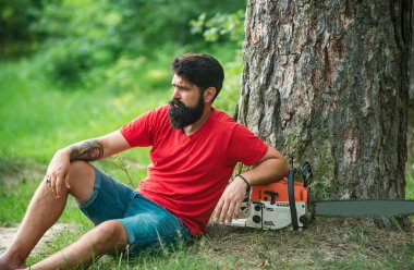 Ormanın yakınında baltası olan yakışıklı bir genç. Elinde elektrikli testereyle oduncu. Elektrikli testere. Ormanların tahrip edilmesi doğal ekosistemlerin bozulması ve istikrarsızlaşmasının başlıca nedenlerinden biridir.