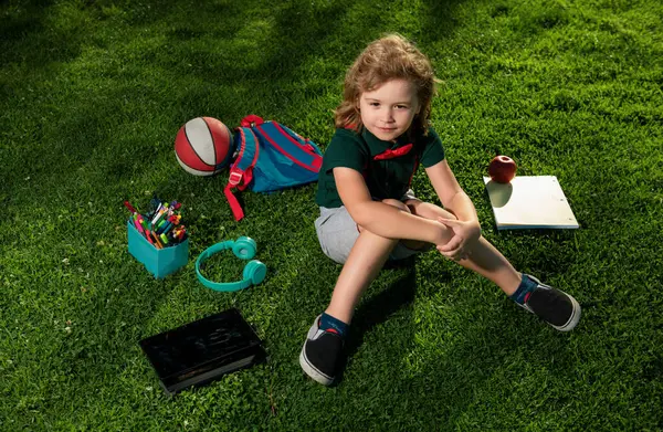 Kid with tablet device and school supplies sitting outdoor in garden, wearing short and shirt. Lttle child outdoor school, education, on line distance learning