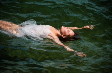 Okyanusta ya da deniz suyunda yüzen güzel genç bir kadın. Rahatlıyor ve kendine bakıyor. Lüks tatil köyünde yaz elbisesi içinde zarif bir kızın moda portresi.