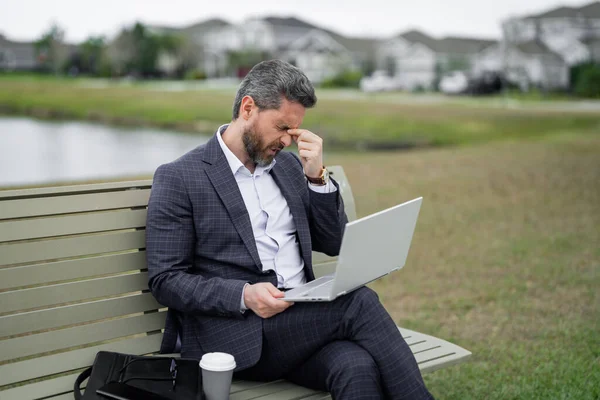 Bored upset tired business man in suit sitting on bench. Tired ceo manager work on laptop. Tired businessman have stress. Head pain migraine. Tired freelancer man online work from laptop in park